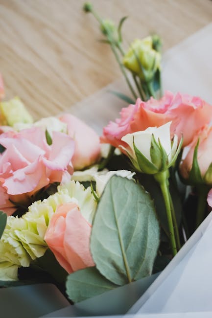 A close-up view of a floral arrangement featuring soft pink and cream lisianthus flowers with delicate, ruffled petals, complemented by fresh green leaves and stems. The bouquet is partially wrapped i