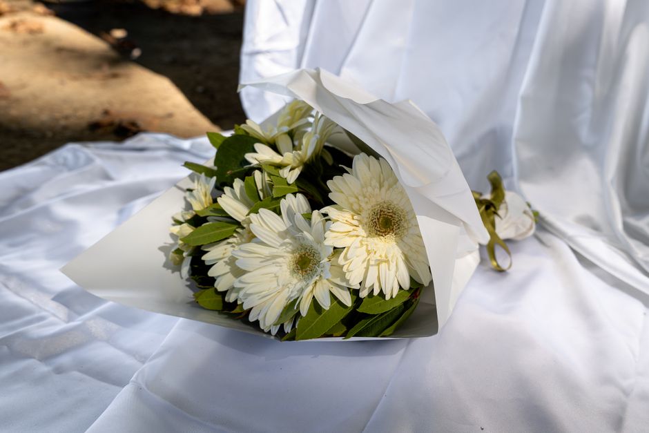 A bouquet of fresh white gerbera daisies and white chrysanthemums with green foliage, wrapped in white paper with a gold ribbon, resting on a satin-like white fabric surface. The flowers are arranged 