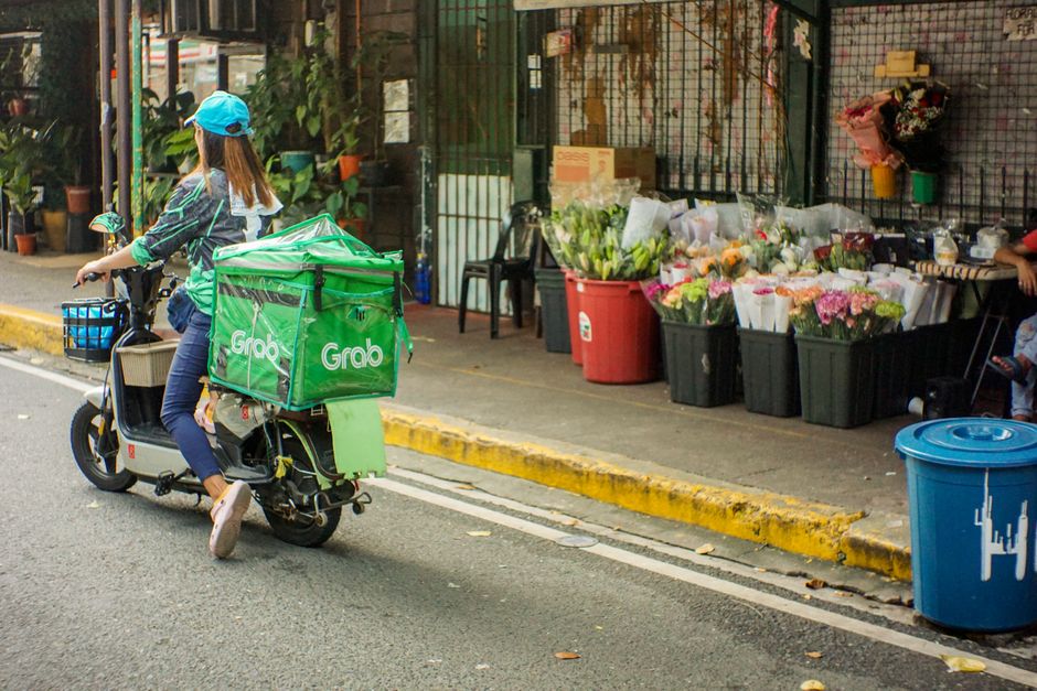 A female delivery rider wearing a blue cap and casual clothing is riding a small electric scooter with a large green Grab delivery box attached to the back, on a street next to a flower shop. The shop