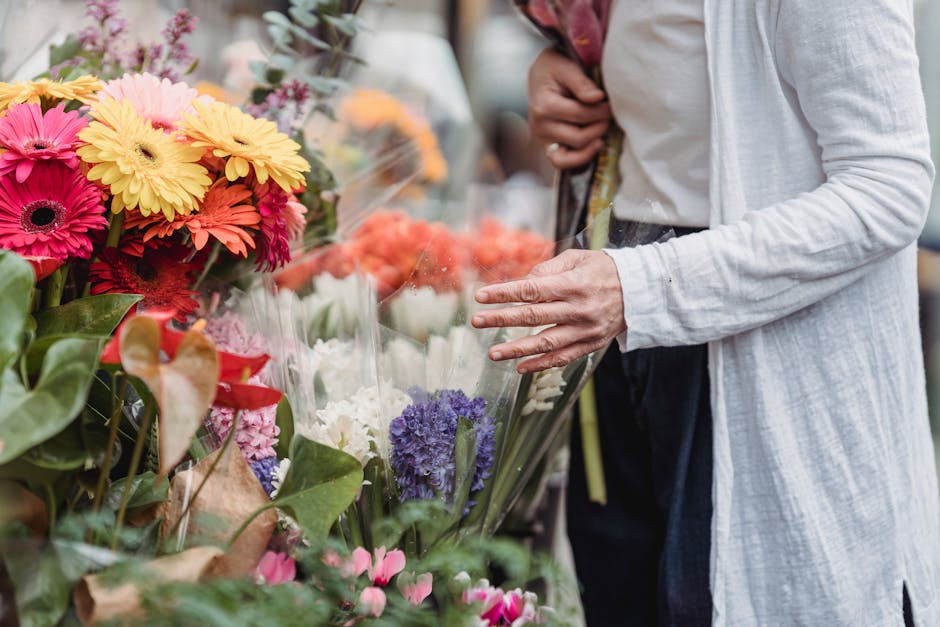 Close-up of a person browsing a vibrant flower stall, with bouquets of fresh, colorful flowers including pink and orange gerbera daisies, purple hyacinths, white blooms, and green foliage. The individ